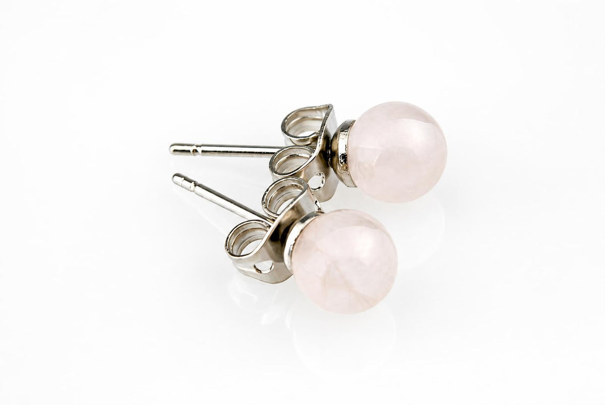 A pair of silver-toned stud earrings with round, pale pink rose quartz stones on a white reflective surface.