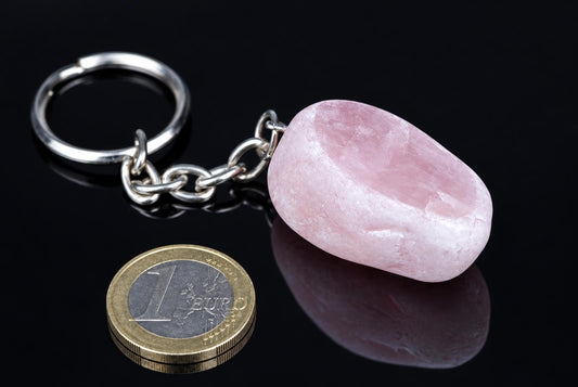 A silver-toned keychain with a smooth, pink stone pendant rests on a dark surface next to a one-euro coin.