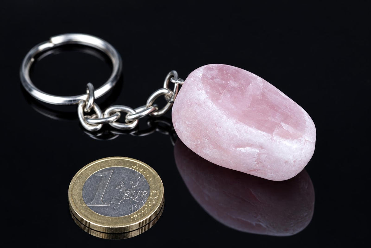 A silver-toned keychain with a smooth, pink stone pendant rests on a dark surface next to a one-euro coin.