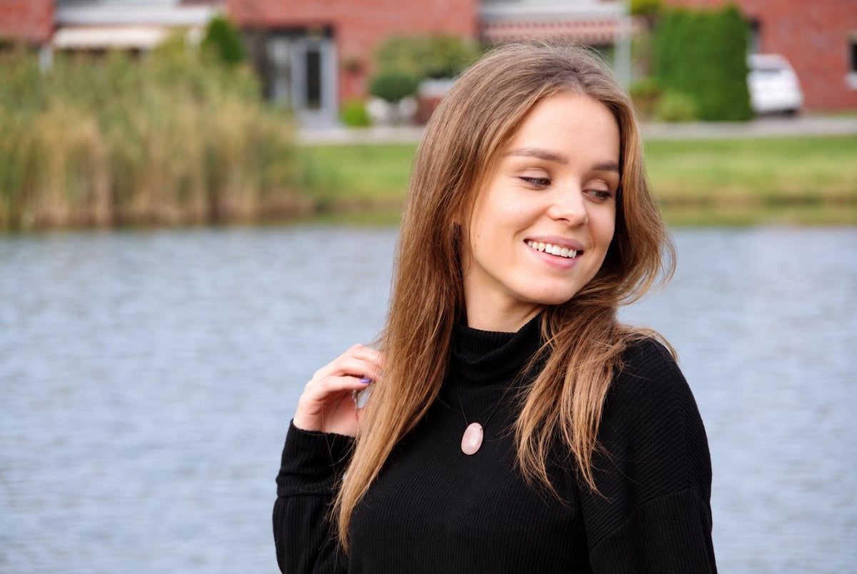 A woman wears a black turtleneck with a pink oval pendant on a dark surface near water.