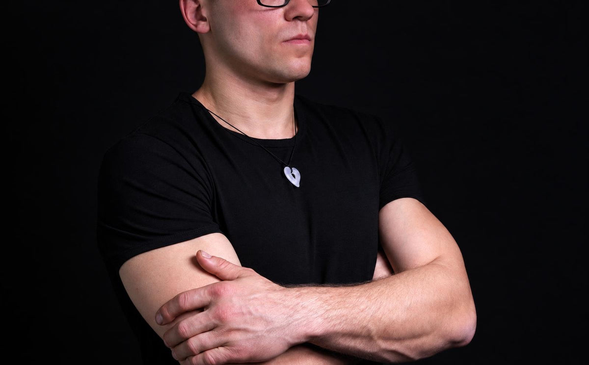 A man in a black t-shirt wears a light-colored heart-shaped pendant on a dark surface.