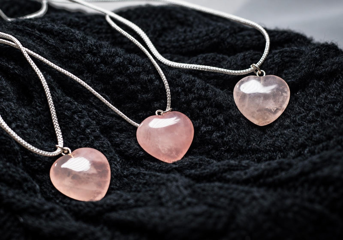 Three silver-toned necklaces with pink heart-shaped rose quartz pendants on a dark knitted surface.