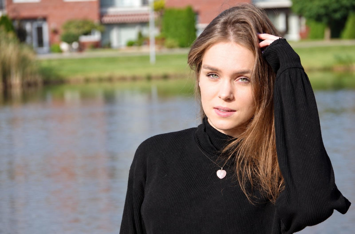 A woman wears a black turtleneck sweater with a small, pale pink heart-shaped pendant on a thin chain.