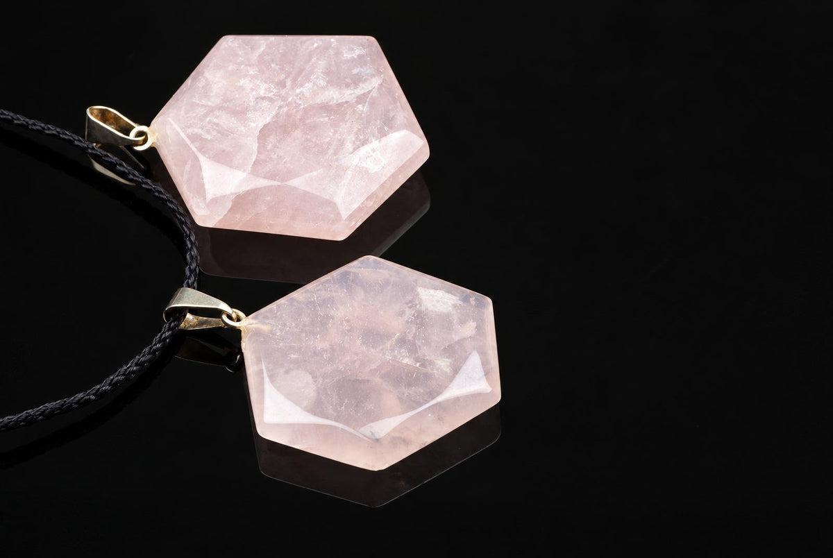 Pale pink hexagonal rose quartz pendants with faceted edges and metal loops lie beside black cords on a glossy dark surface.