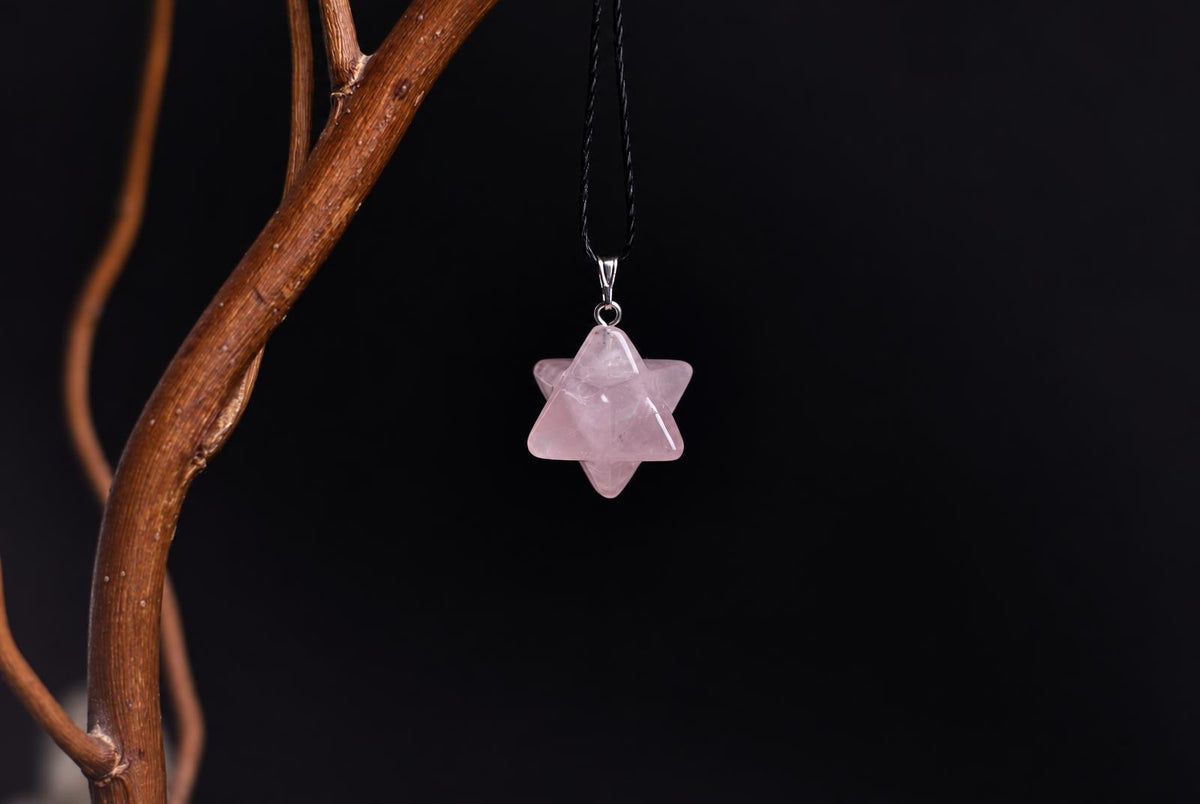 A pink star-shaped pendant hangs from a black cord against a dark background, with a brown branch visible on the left.