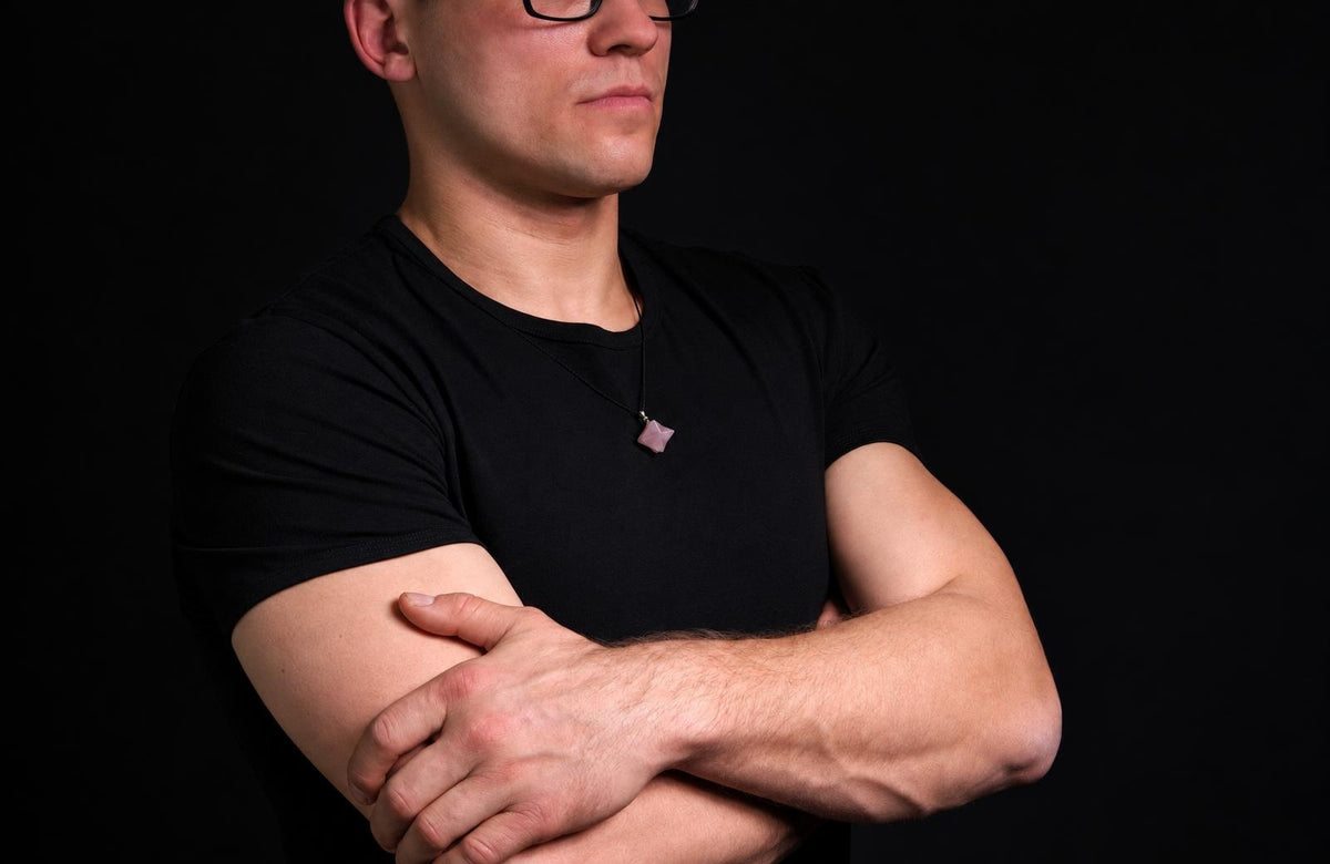 A man in a black t-shirt wears a necklace with a small, pale pink pendant against a dark background.