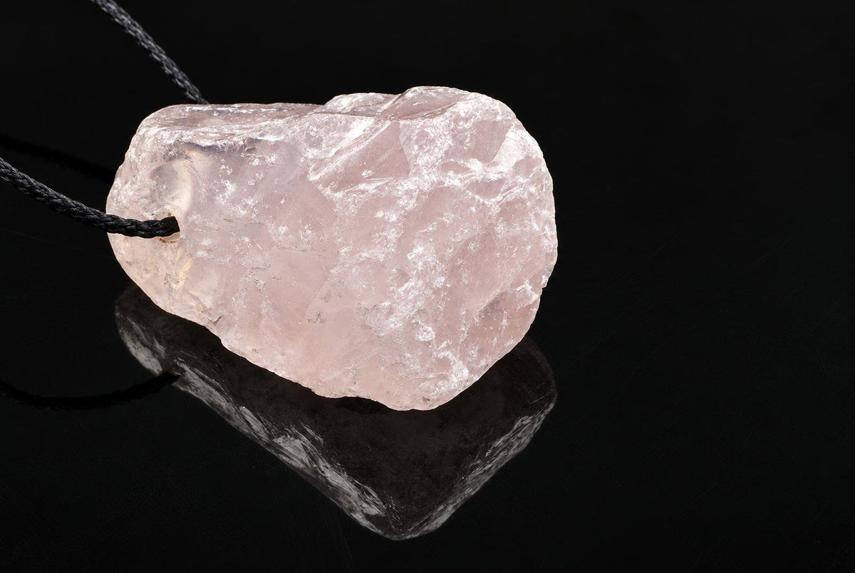 A pale pink, translucent rose quartz pendant with a rough natural shape hangs on a black cord against a dark reflective surface.