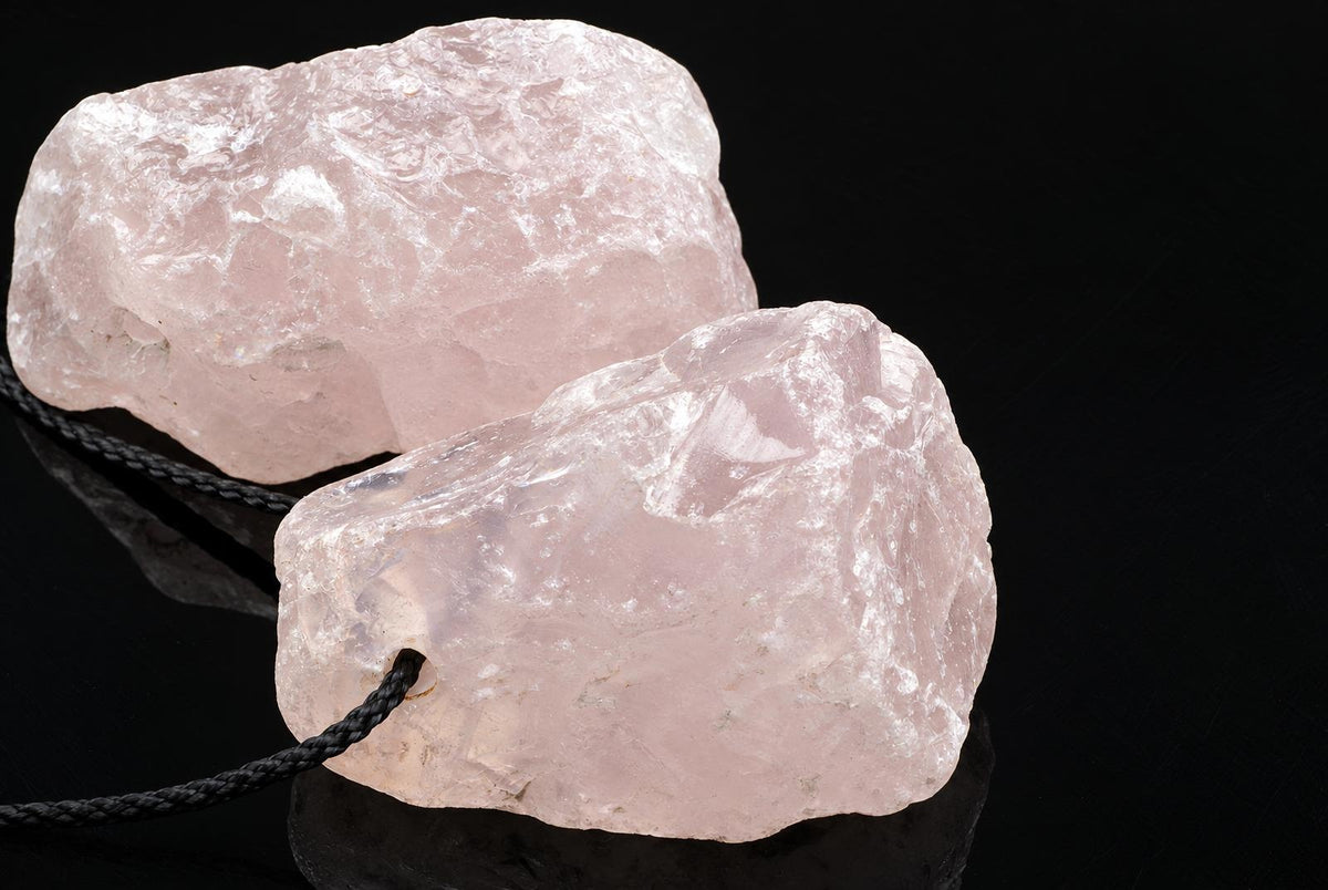 Two pale pink rose quartz stones on a dark surface, one with a black cord looped through it.