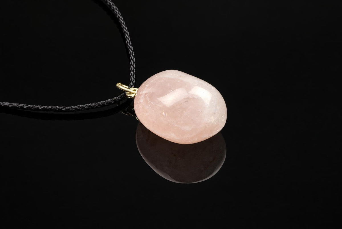 A smooth, oval-shaped pink stone pendant on a dark cord with gold-toned metal against a black reflective surface.