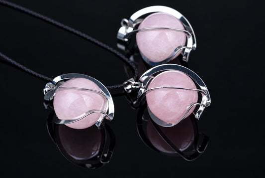 Three silver-toned pendants with pink spherical stones on dark cords, arranged on a reflective black surface.