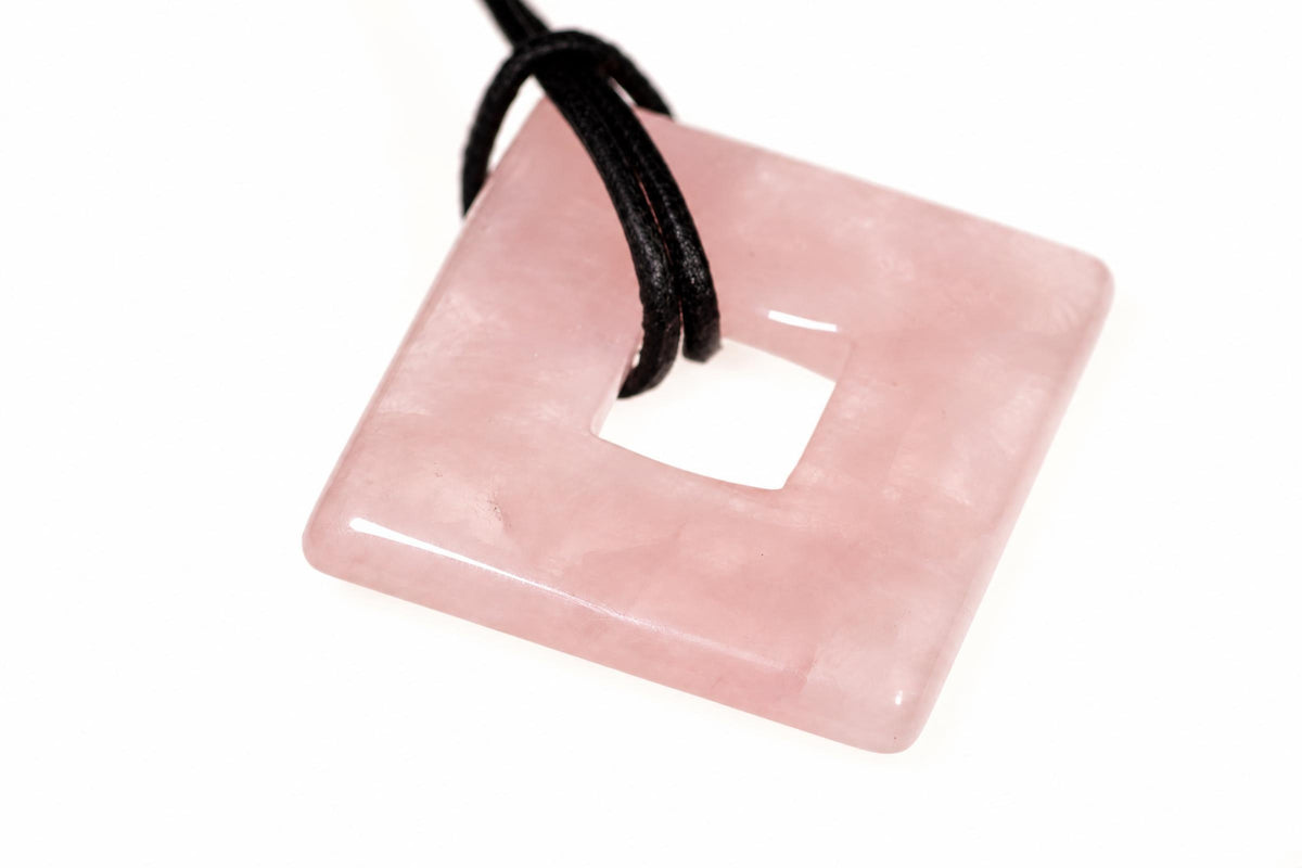 A square, pink rose quartz pendant with a central hole and a black cord looped through it, set against a white background.