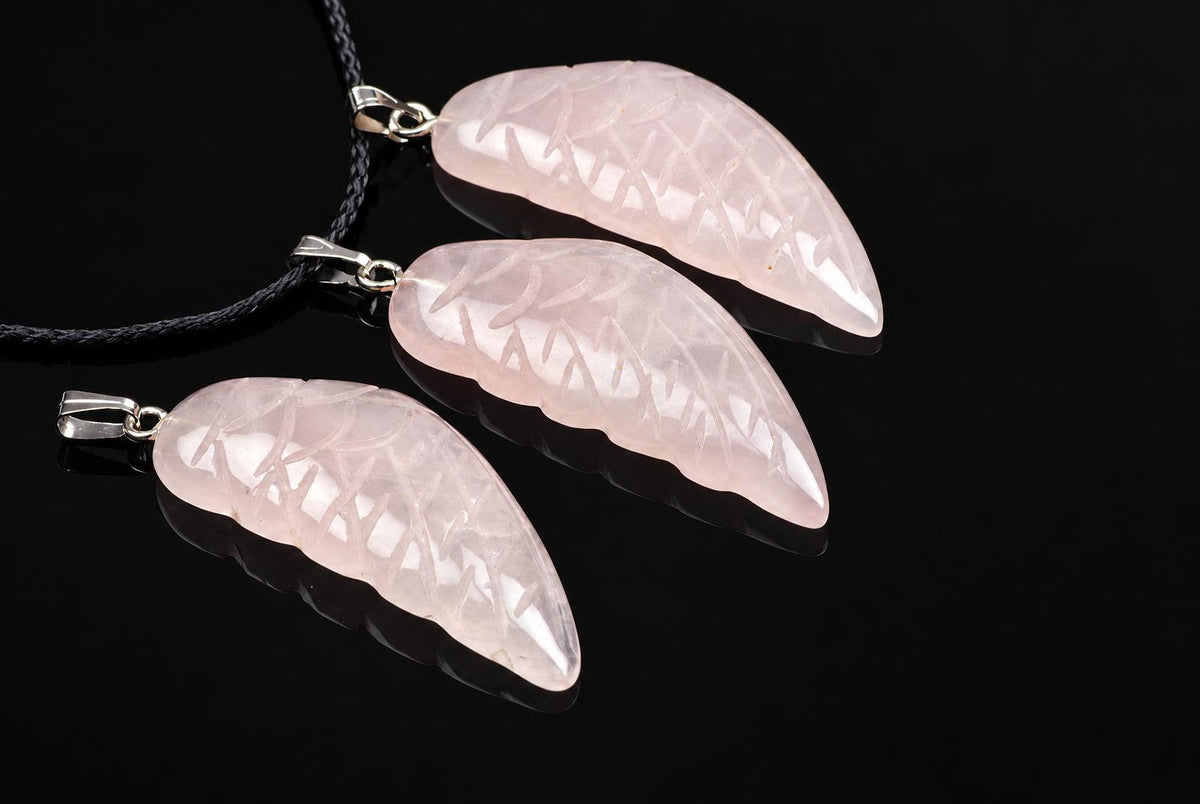 Pale pink rose quartz wing pendants with carved feather grooves and silver-tone loops lie beside black cords on a glossy dark surface.