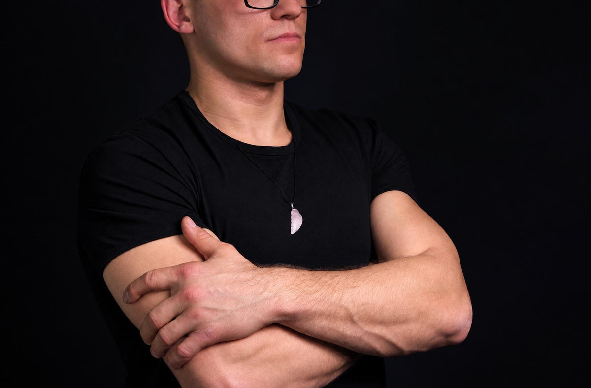 A man in a black t-shirt wears a pale pink wing-shaped pendant on a dark surface.
