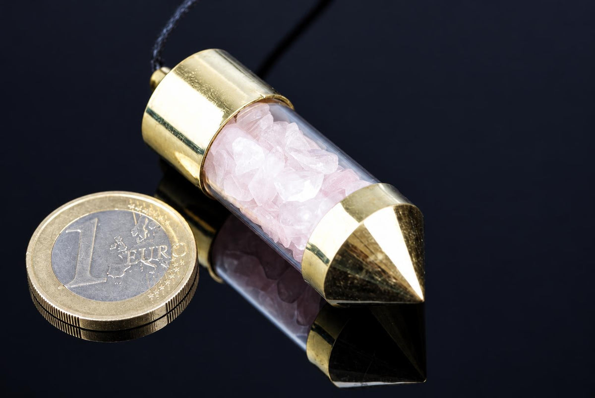 A brass pendulum with a clear tube containing pink rose quartz crystals, resting on a dark reflective surface next to a one-euro coin.
