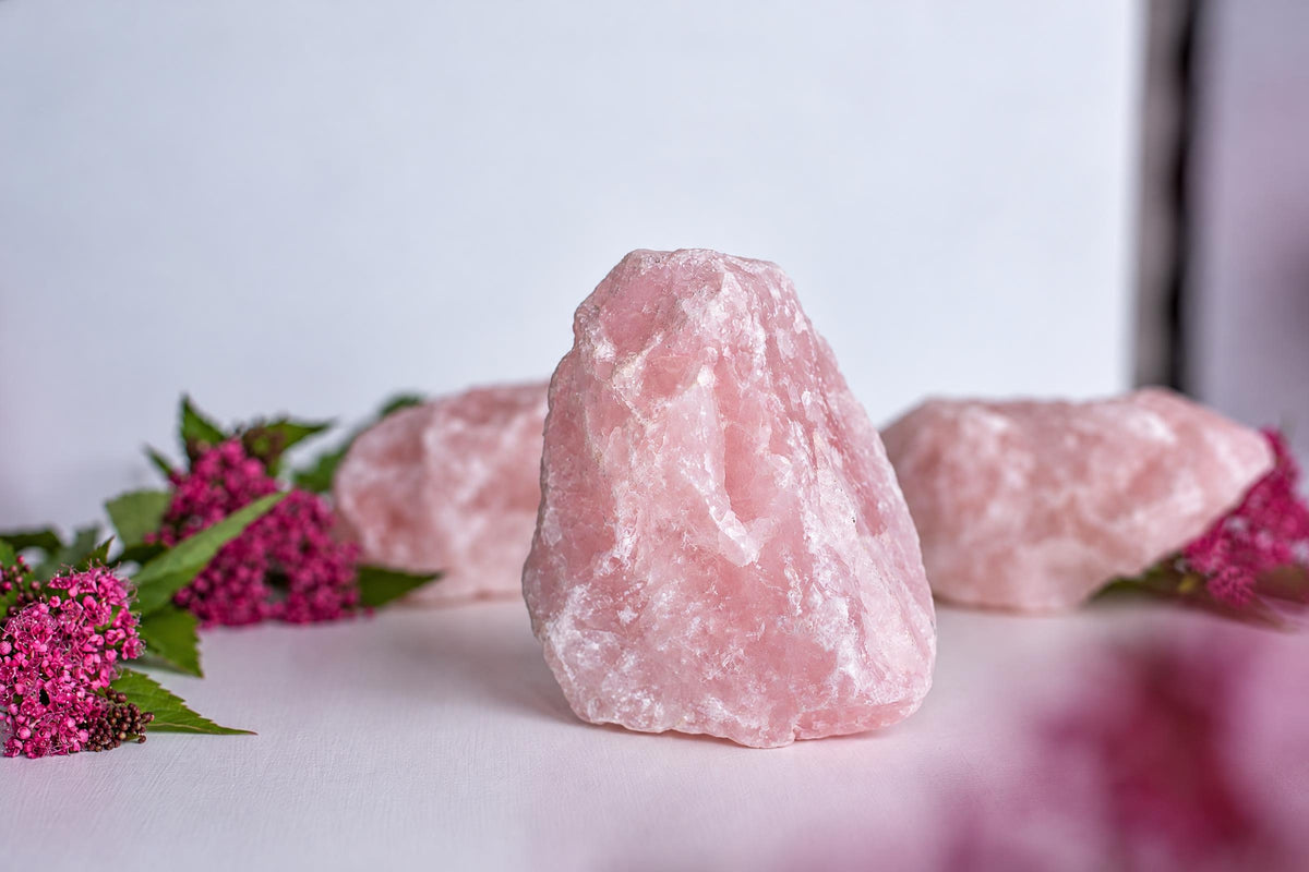 Three pink rose quartz crystals on a pale surface with pink flowers nearby.