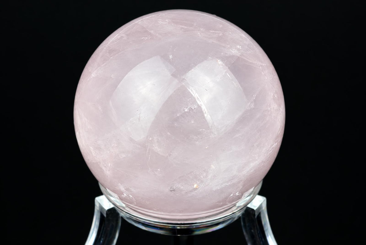 A polished pink rose quartz sphere rests on a clear stand against a dark background.