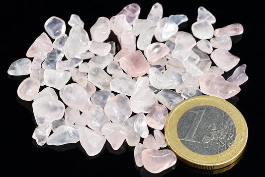 A pile of small, translucent pink and white rose quartz tumbled stones on a dark surface next to a one euro coin.
