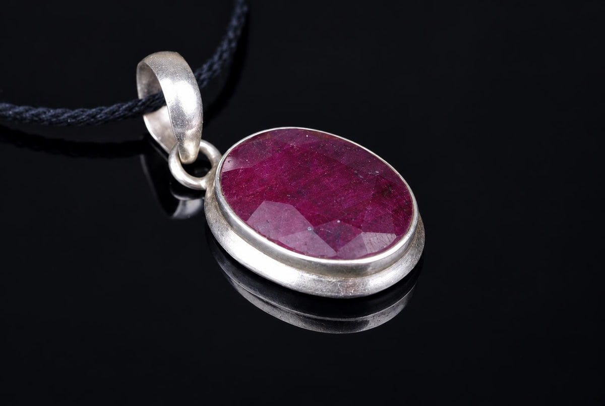 A silver-toned pendant with a faceted, oval red stone on a dark cord, resting on a black reflective surface.