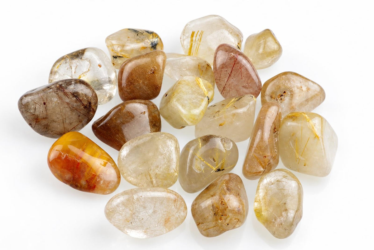 A pile of polished rutile quartz stones in brown, yellow and clear tones with golden needle-like inclusions on a white surface.
