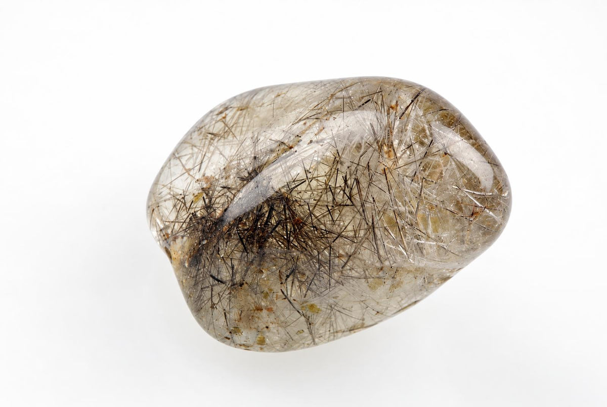 A polished, translucent Rutile quartz stone with a smooth, rounded shape. It contains numerous dark brown and black needle-like inclusions set against a pale, clear background on a white surface.