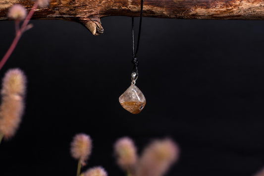 A clear, teardrop-shaped pendant with golden rutile inclusions hangs from a black cord on a dark surface.