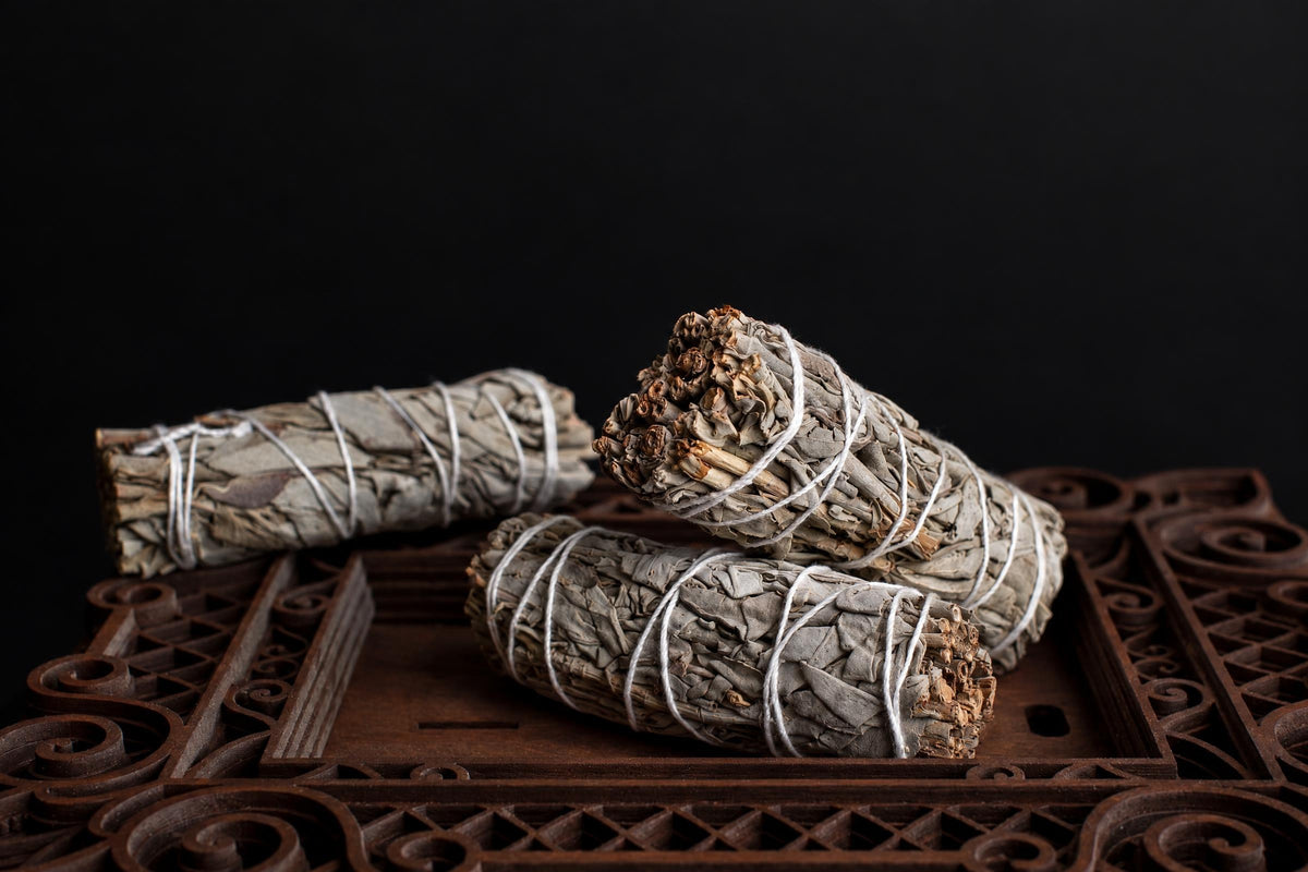 Three dried sage bundles tied with white string rest on a dark wooden surface against a black background.