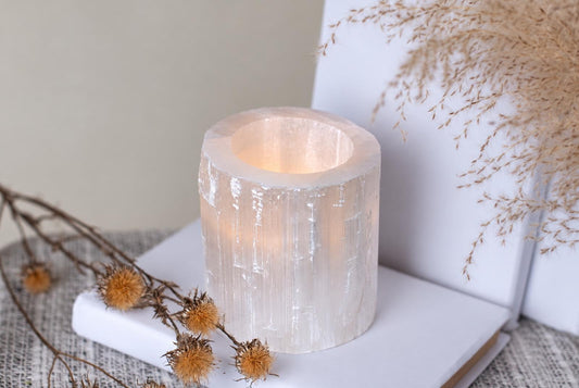 A cylindrical, translucent white selenite candle holder with a warm glow inside, resting on a white book and pale surface.