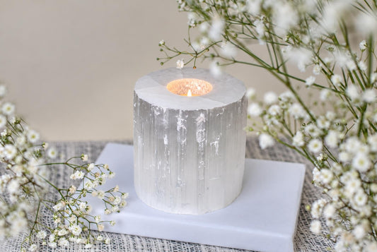 A cylindrical selenite candle holder with a lit tea light inside, resting on a white square base surrounded by baby's breath flowers.