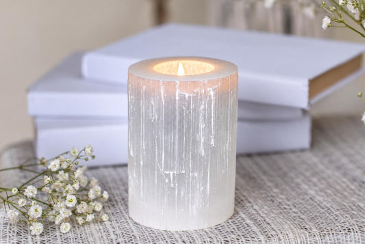 A lit candle inside a translucent, white cylindrical holder with vertical striations sits on a pale woven surface.