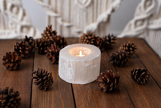 A lit candle inside a white, cylindrical selenite holder sits on a dark wooden surface surrounded by brown pinecones.