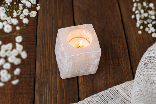 A square, pale pink selenite candle holder with a lit tea light inside, on a dark wooden surface.
