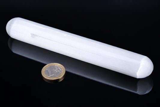 A white, cylindrical selenite massage stick with rounded ends rests on a dark surface next to a one-euro coin.