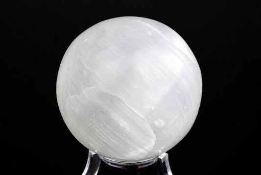 A polished white selenite sphere with visible striations sits on a clear stand against a dark background.
