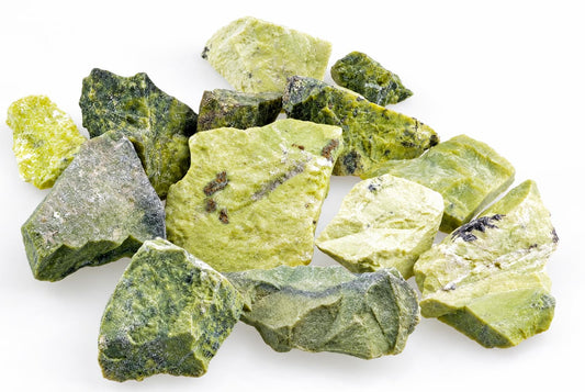 A pile of raw Serpentinite stones in various shades of green, some with dark specks and white patches, on a pale surface.