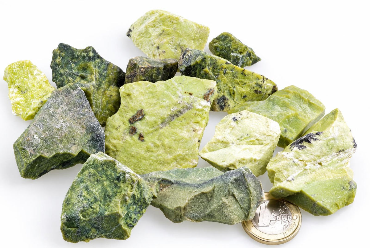 A pile of raw, irregular green serpentine mineral pieces on a white surface with a one-euro coin for scale.