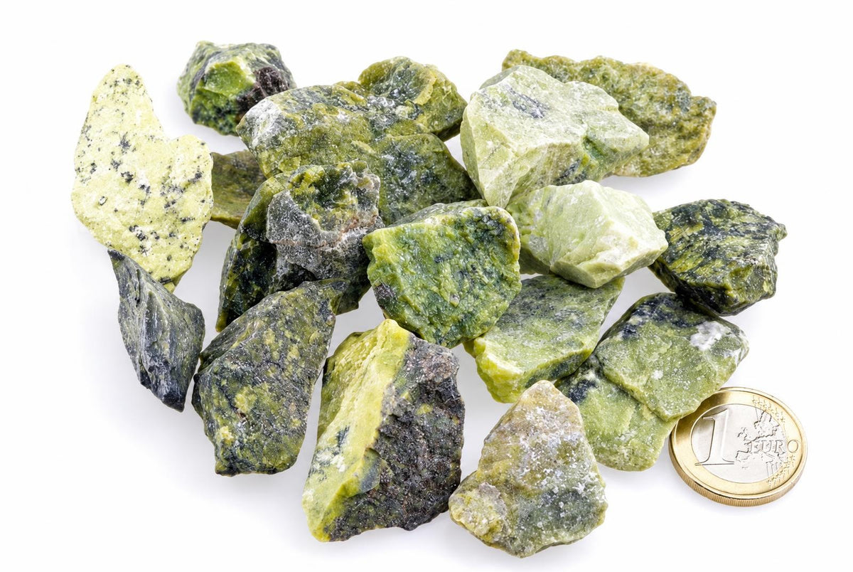 A pile of raw, irregular green and yellow-green mineral chunks with dark inclusions sits on a white surface next to a one-euro coin.
