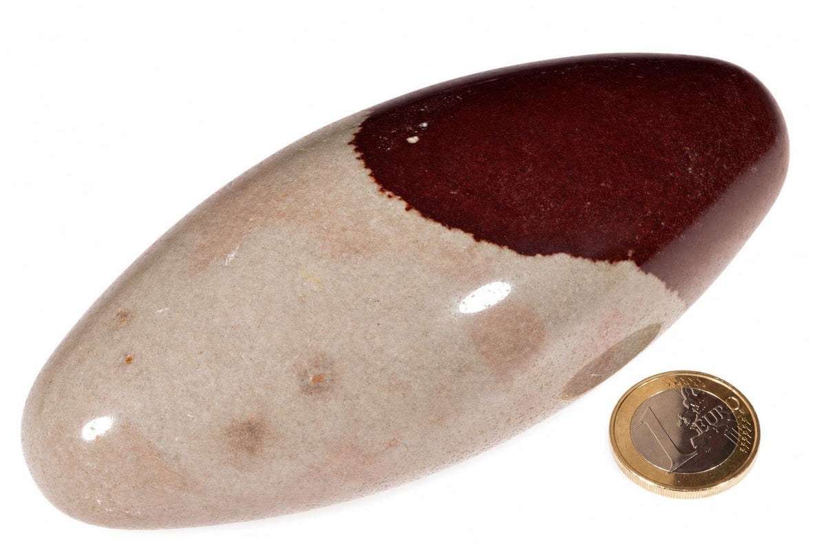 A polished, smooth stone with a light beige base and a dark reddish-brown upper section, shown next to a one-euro coin on a white surface.