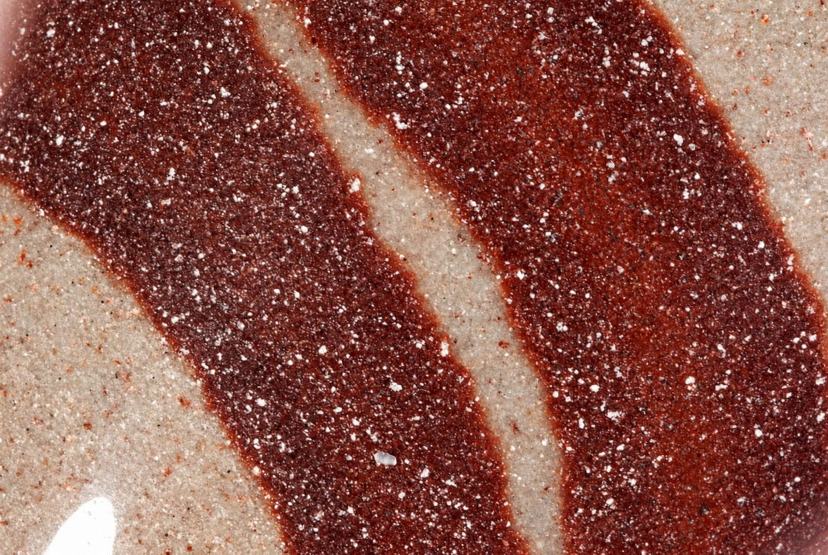 A close-up of a polished surface with dark reddish-brown diagonal stripes on a pale background, sprinkled with white granules.