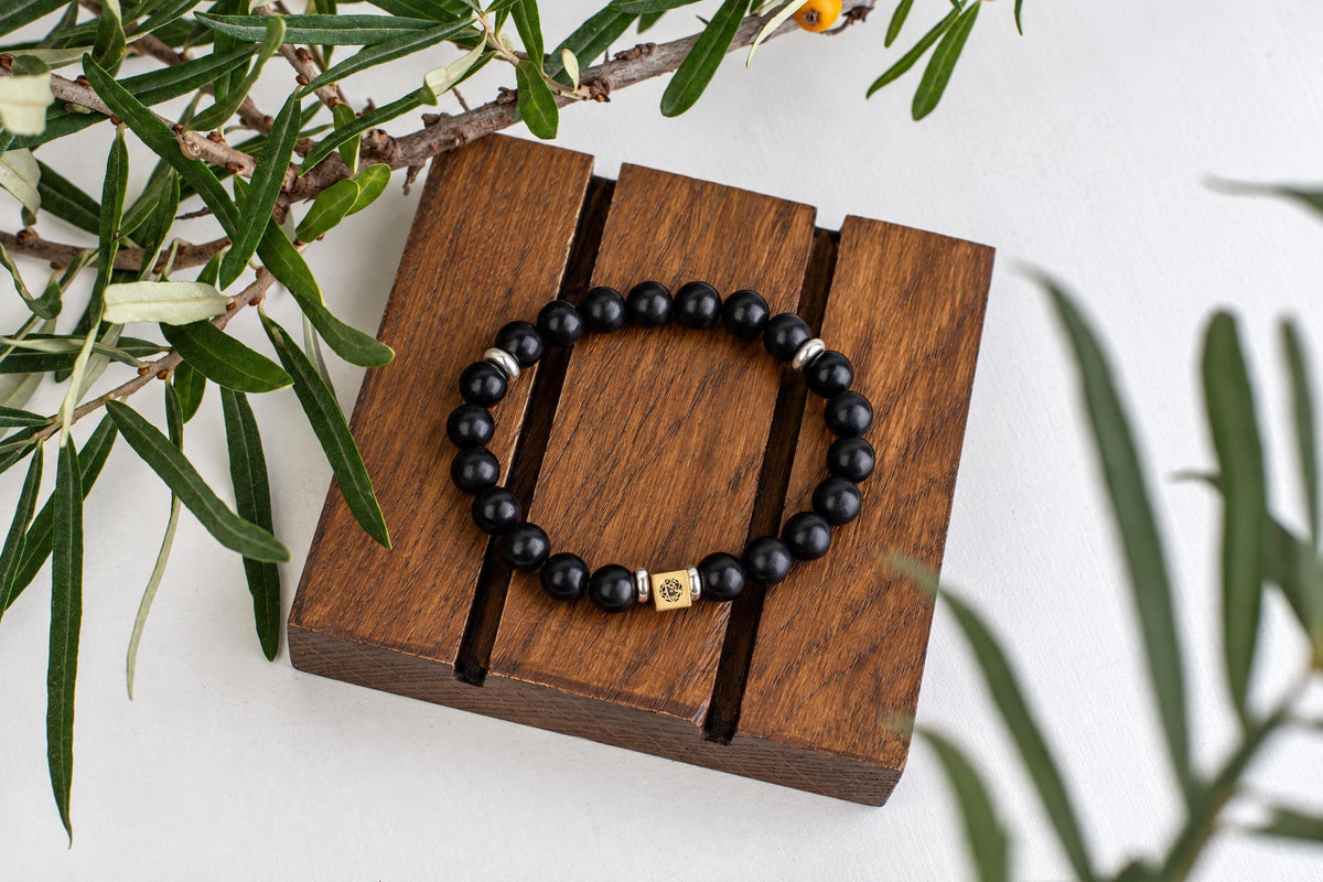 A bracelet of black round beads with a small square metal bead rests on wooden slats beneath narrow green leaves.