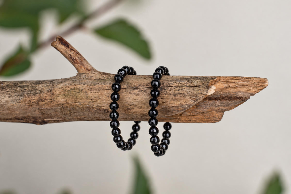 Two black faceted bead bracelets rest on a rough wooden branch with blurred green leaves in the background.
