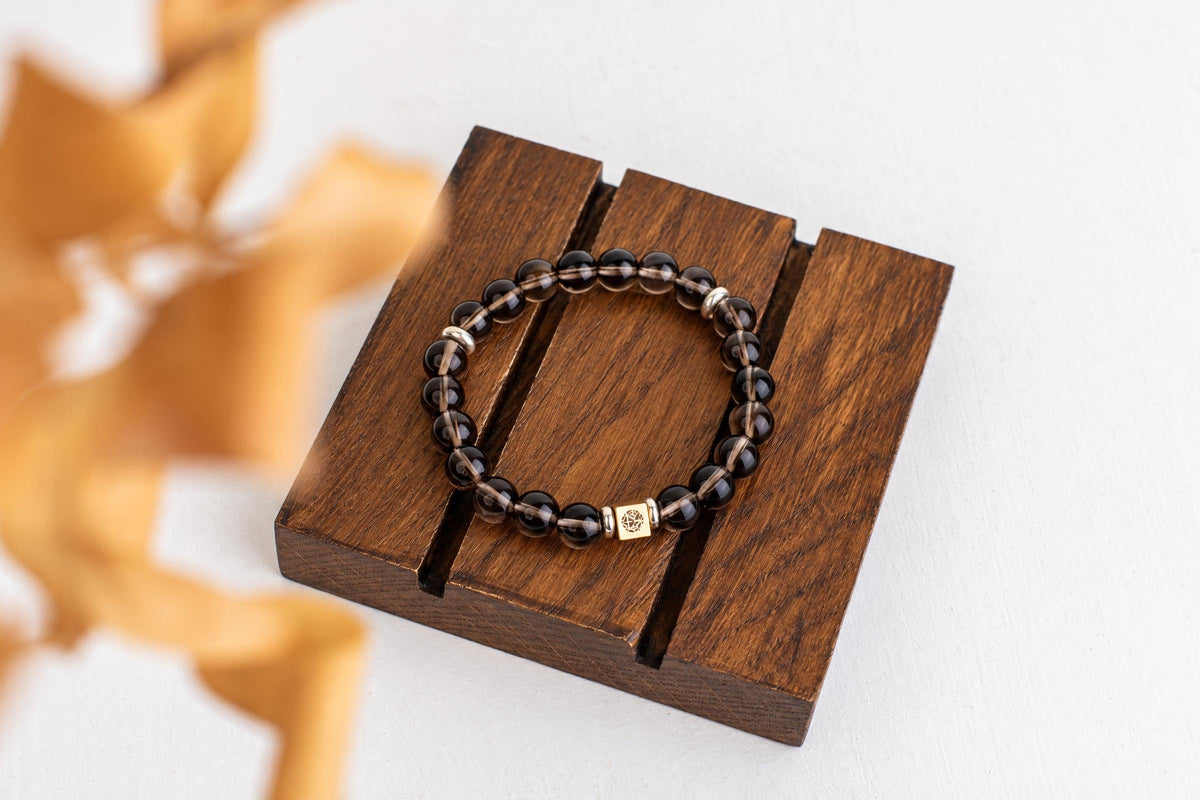 A bracelet with dark, translucent beads and a gold-toned square charm rests on a wooden display stand.