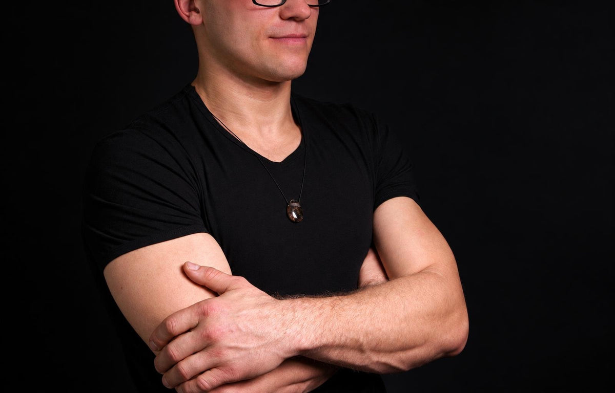 A man in a black t-shirt wears a dark brown, translucent pendant on a thin cord against a dark background.
