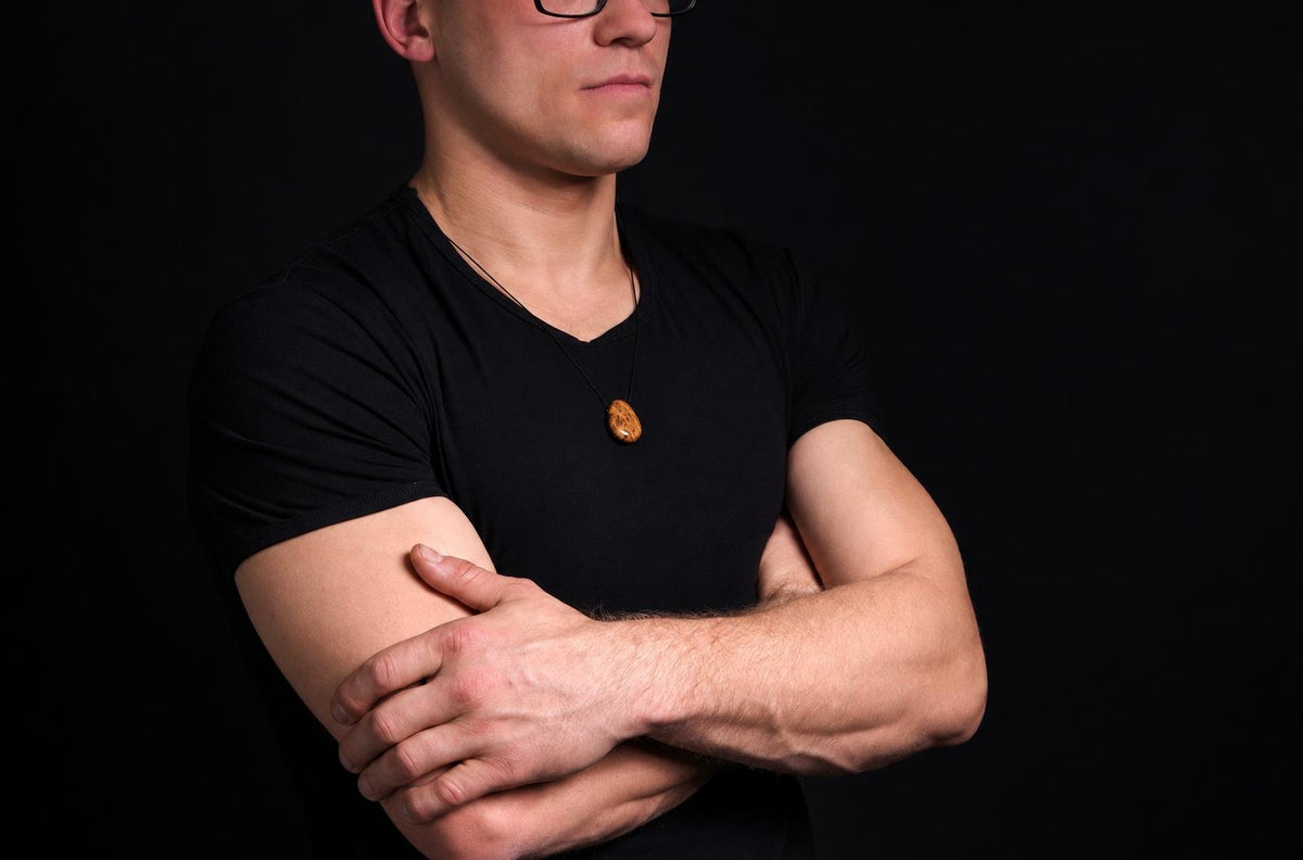 A man in a black shirt wears a dark brown oval pendant on a thin cord against a dark background.
