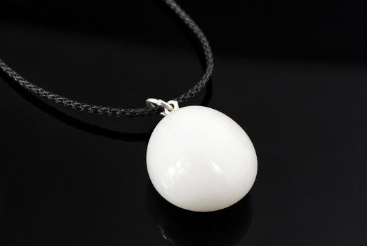 A smooth white snow quartz pebble pendant hangs from a black cord, with a soft reflection on the dark surface below.