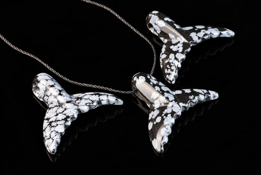 Three dolphin tail pendants with white snowflake patterns on dark stone, attached to twisted cords, arranged on a black reflective surface.