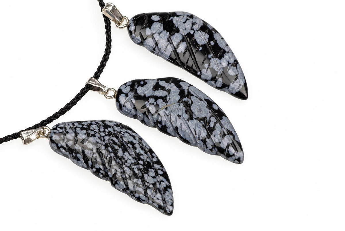 Three black and white snowflake obsidian pendants shaped like wings hang from a dark cord on a pale surface.
