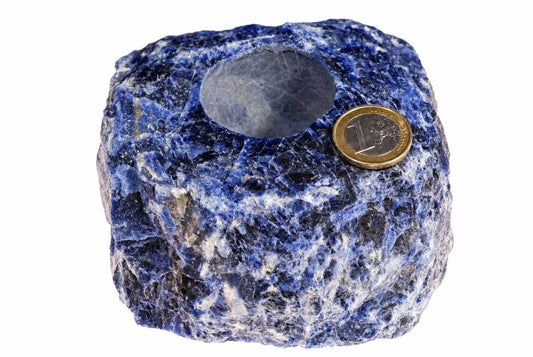 A rough, blue and white sodalite stone with a hollowed-out top opening, shown on a white surface next to a one-euro coin.