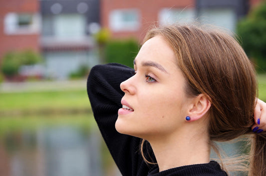 A woman wears a small, round dark blue stud earring in her earlobe against an outdoor background.
