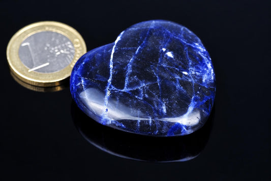 A polished, heart-shaped sodalite stone with deep blue and white veining rests on a dark surface next to a one-euro coin.