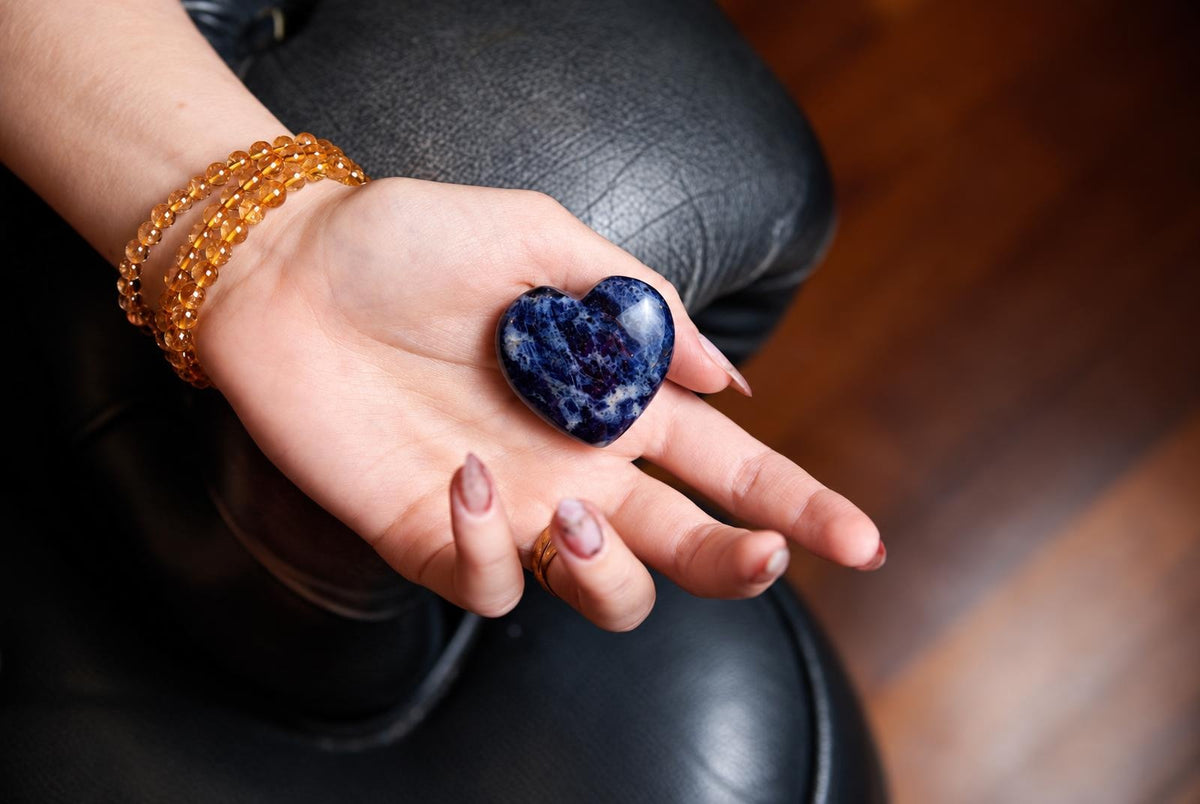 A hand holds a polished, heart-shaped sodalite stone with deep blue and white patterns on a dark surface.